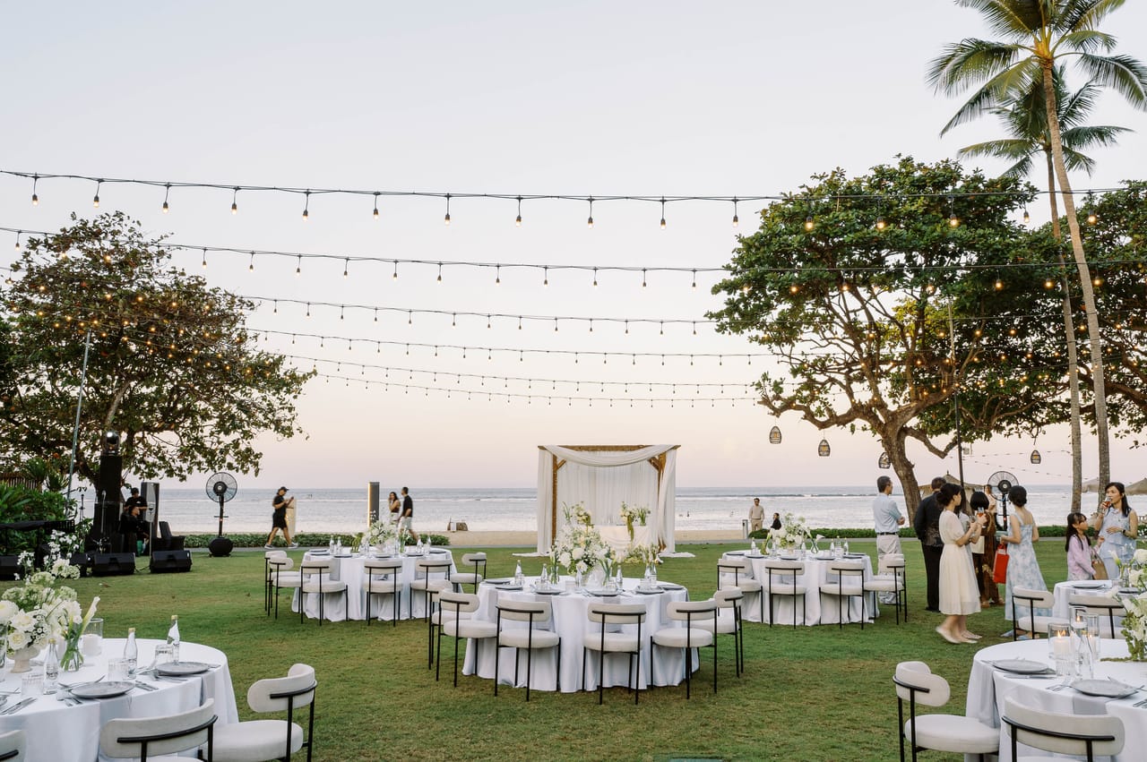 An oceanfront Bali wedding reception with white tables beneath string lights.