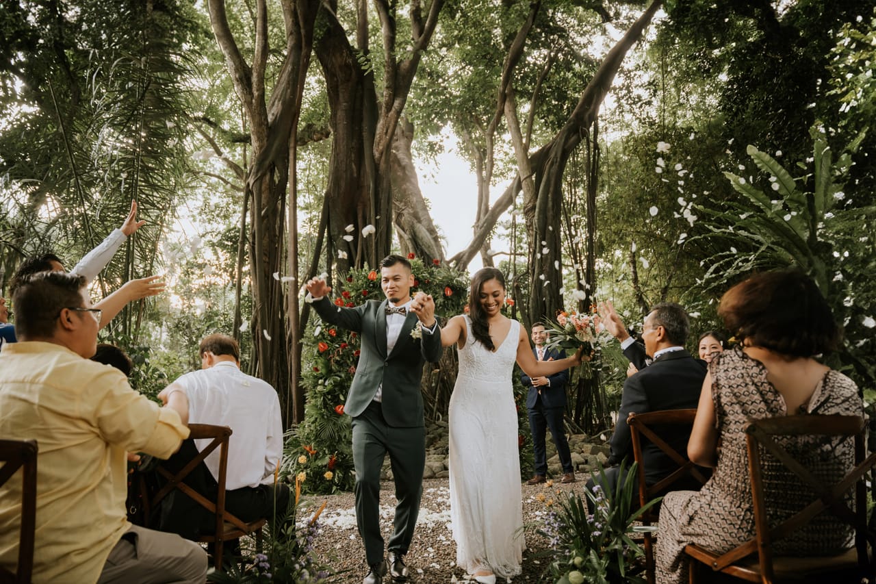 Alexandra and Raymond walking from their ceremony beneath tropical trees and red flowers